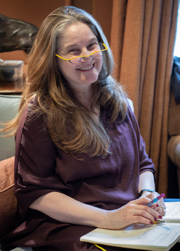 Kate Hammer, a middle-aged white woman wearing yellow half-spectacles smiles. Her burgundy tunic hangs loose. On her lap, a notebook. She holds a pen. Her nails are painted merlot.