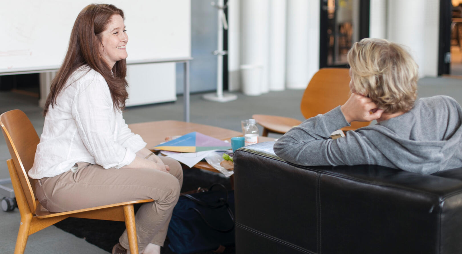 Kate Hammer, a white middle aged woman wearing a linen shirt and tan chino trousers in side profile listening and smiling towards a seated workshop participant at SoPact in 2015.